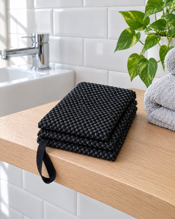 La serviette Seshin noire, pliée sur une tablette en chêne dans une salle de bain blanche ; une de ses poignées pend.