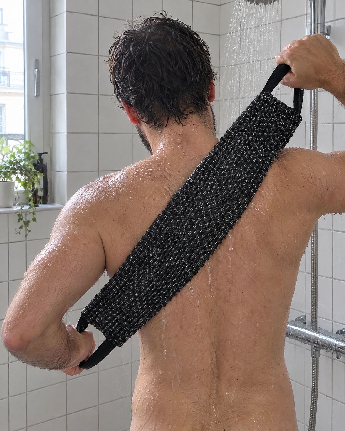 Un homme de dos sous la douche tend la serviette Seshin noire en diagonale dans son dos, en la tenant par ses deux poignées.